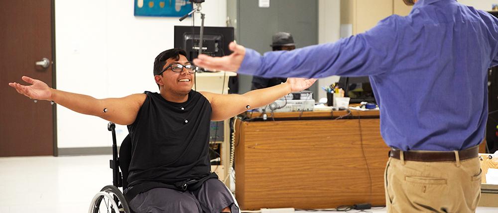 Two men in a lab, one in a wheel, face each other smiling.